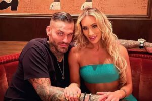 A tattooed man and woman sit close together on a red booth, holding hands and smiling at the camera in a cafe setting.