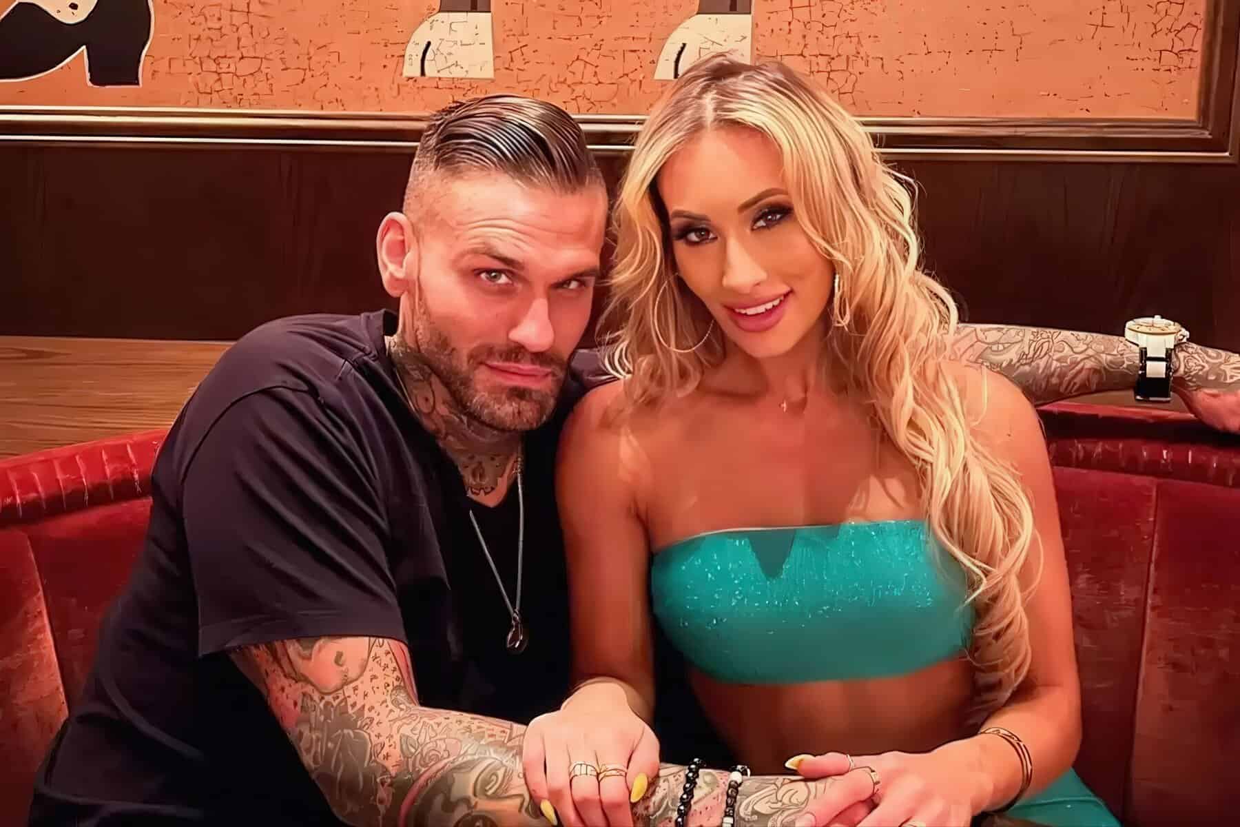 A tattooed man and woman sit close together on a red booth, holding hands and smiling at the camera in a cafe setting.