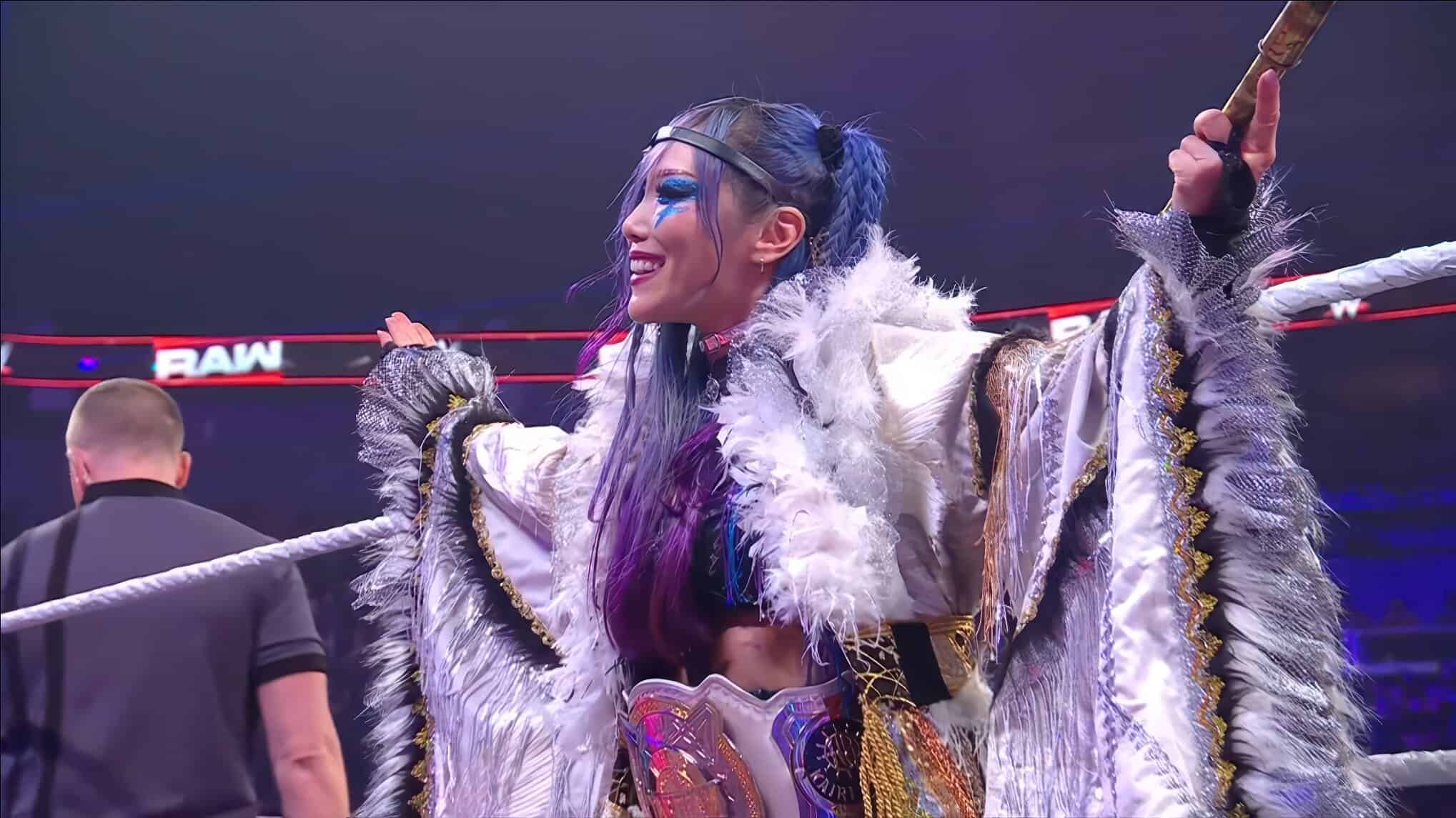Female wrestler in a silver, feathered costume raises arms in a wrestling ring, blue face paint and hair visible, crowd in background.