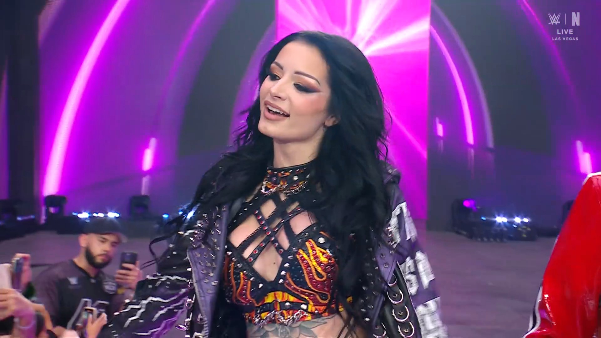 Female wrestler/performer on a neon-lit stage, wearing a black studded top with flame design and a leather jacket; audience films.