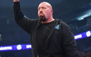 Bald male wrestler in a black hoodie raises his right arm in a wrestling arena.
