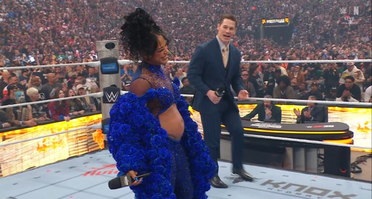 Pregnant woman in a blue rose-adorned gown smiles in a WWE ring, holding a microphone as a host or performer speaks to a cheering crowd.