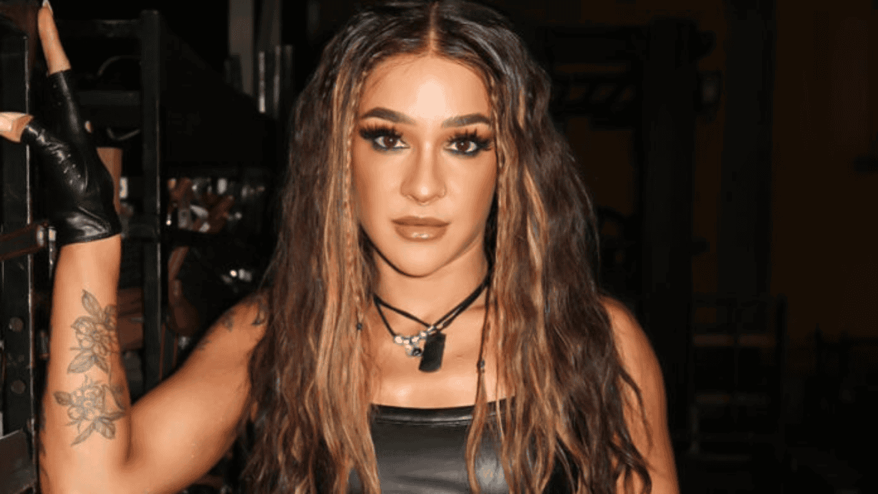 Person with long brown hair and bold makeup wearing a black leather top, posing with a tattooed arm visible in a dim backstage setting.