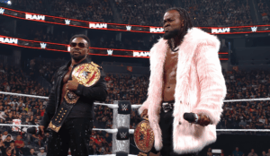 Two WWE wrestlers pose in the ring with championship belts, one wearing a black outfit and sunglasses, the other in a pink fur coat as the crowd roars in the background.